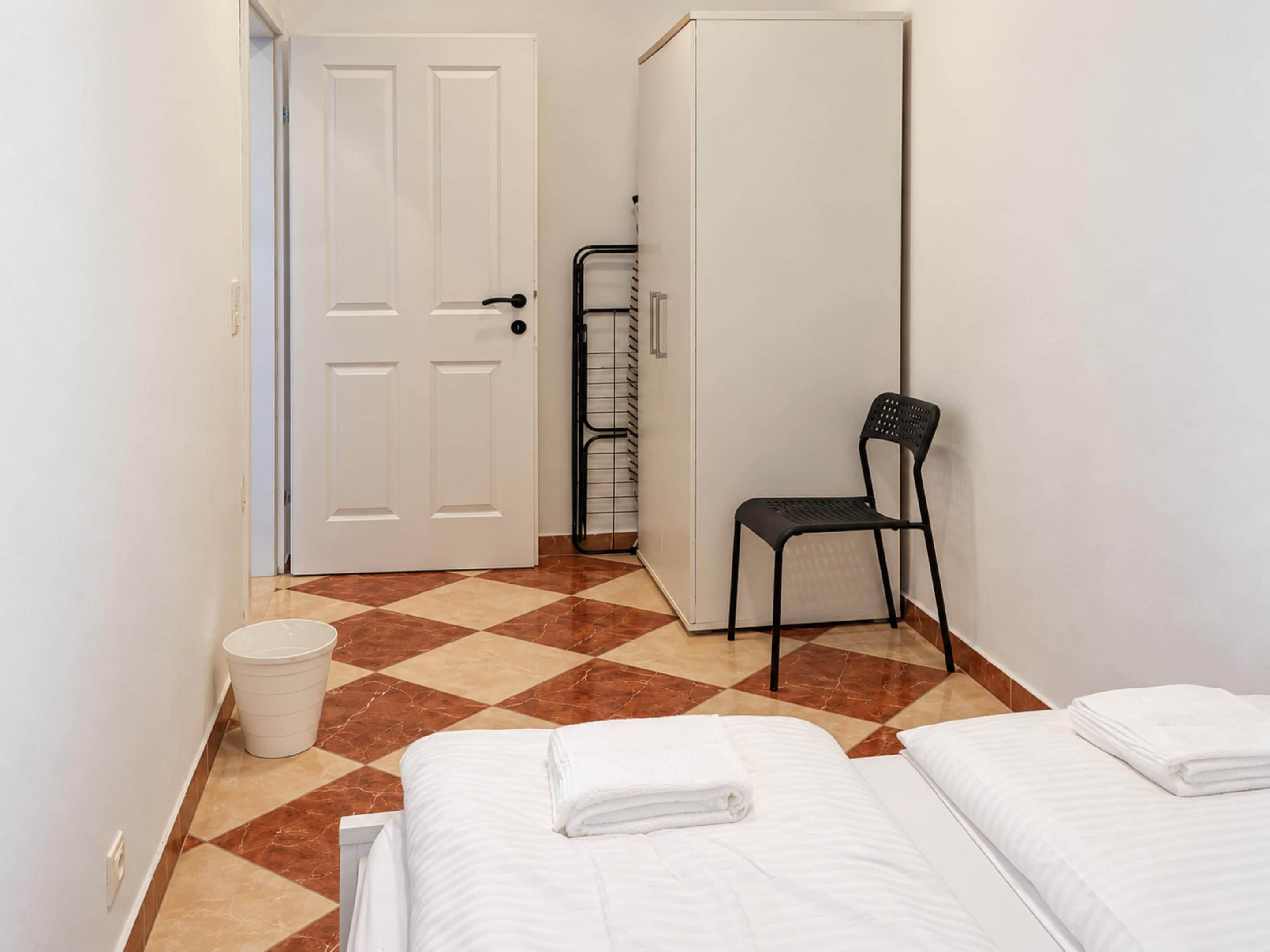 A hotel room with a checkered floor, a bed with a white comforter, a chair, a wardrobe, and a door.