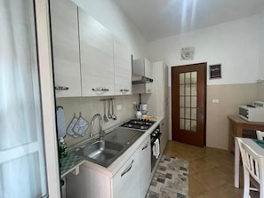 A compact kitchen with white cabinets, a stainless steel sink, and a stove. There's a wooden door with a glass panel and a small table with a chair. - Yes Rome Holidays - One Bedroom Apartment, Sleeps 5 (Rome)