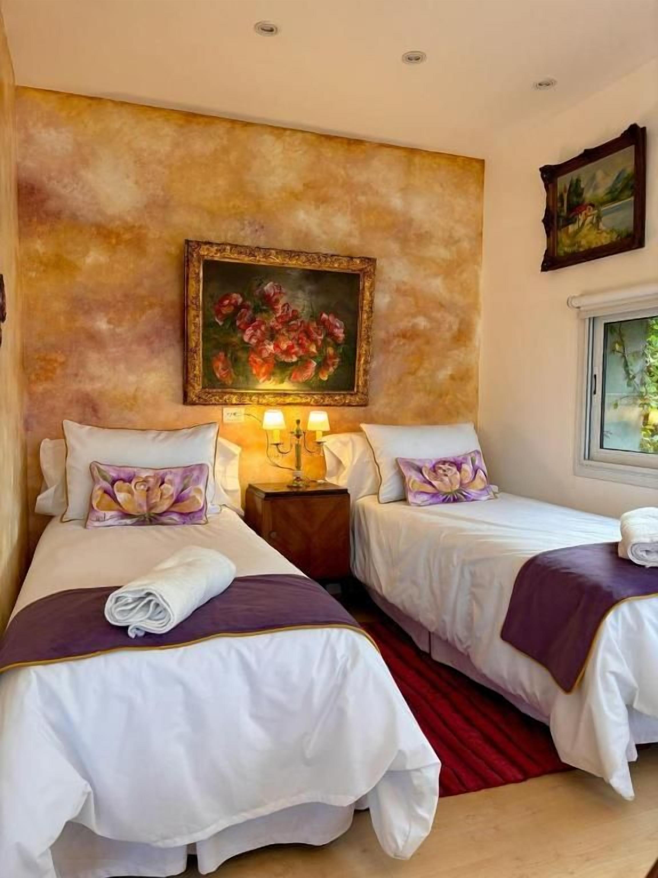 A hotel room with two beds, a painting on the wall, and a window with a view.