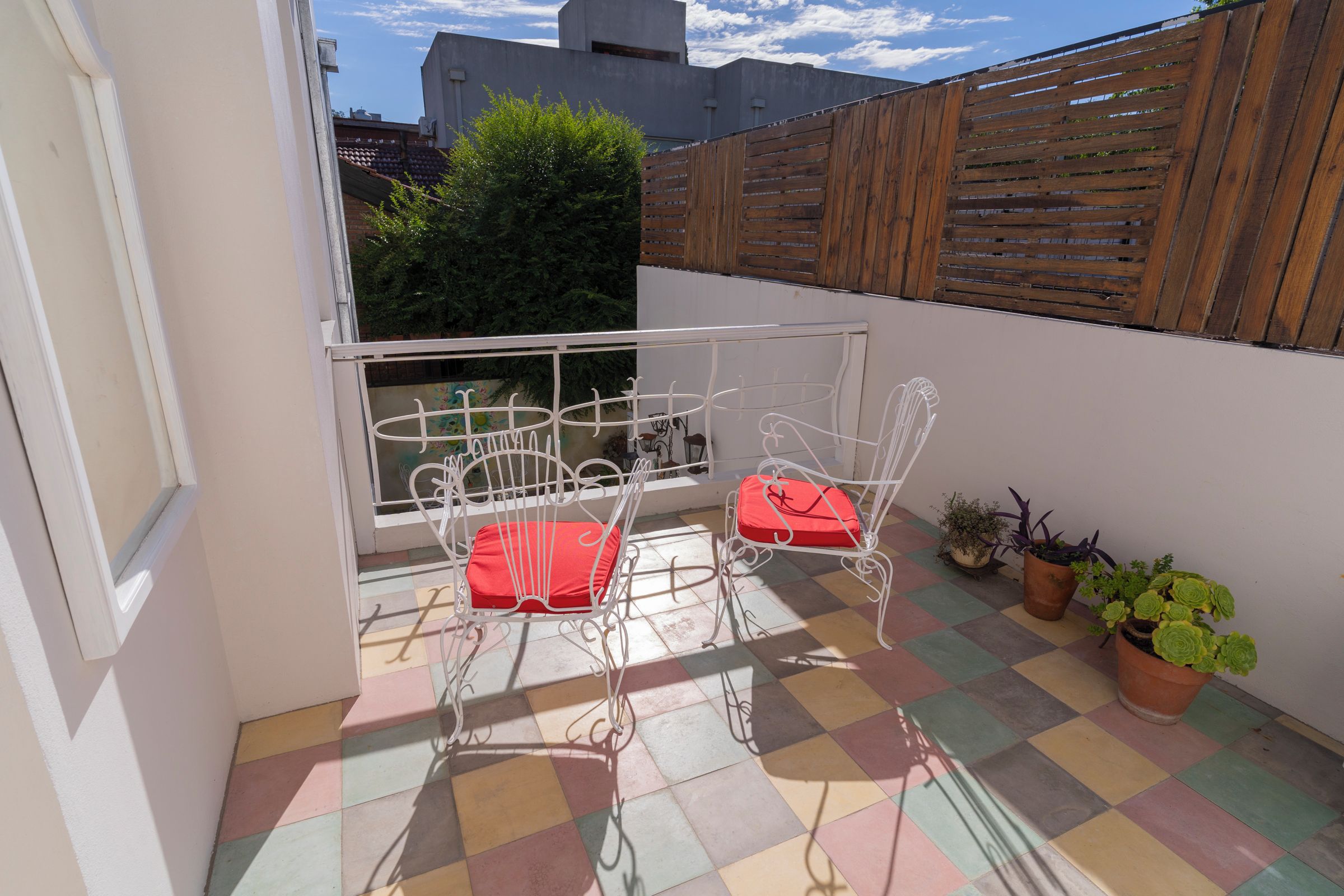 A small balcony with a checkered floor, two white metal chairs with red cushions, and potted plants.