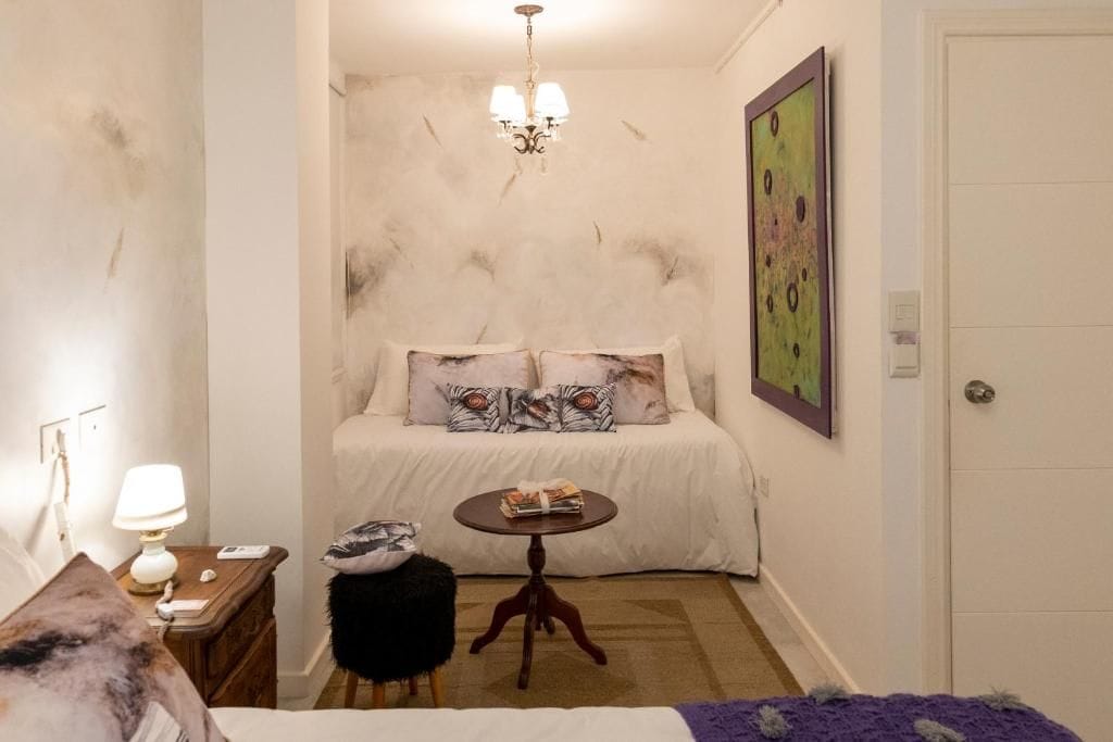 A bedroom with a bed, a small round table, a chair, a lamp, and a painting on the wall.