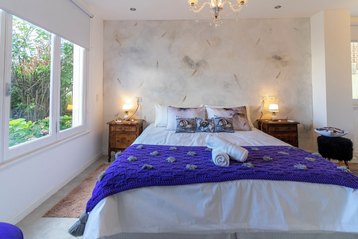 Een slaapkamer met een groot bed, twee nachtkastjes met lampen, een kroonluchter en uitzicht op groen buiten het raam.