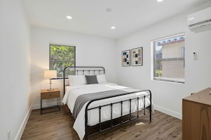 A bedroom with a bed, bedside table, lamp, window with blinds, and a wall-mounted air conditioner. - 1946 Pierce Street (Hollywood)