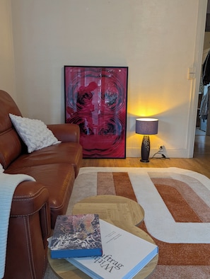 Ein Wohnzimmer mit braunem Ledersofa, einem weißen Zierkissen, einem farbenfrohen abstrakten Gemälde, einer Lampe und einem Teppich.