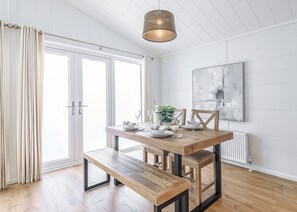 A modern dining area with a wooden table, bench, and pendant lighting. - The Glades Lodges with Hot Tubs (Westbury)