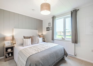 A bedroom with a bed, bedside lamps, a window with curtains, and a wall-mounted light fixture. - The Glades Lodges with Hot Tubs (Westbury)