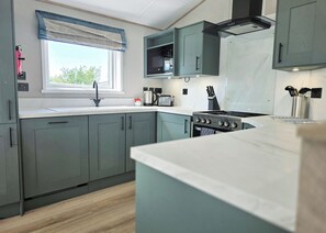 A modern kitchen with grey cabinets, a stainless steel stove, a microwave, and a window with a blue valance. - Platinum 8 Berth Lodge (Mablethorpe)