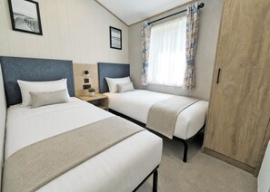 A twin room with two beds, a nightstand, a potted plant, and a window with curtains. - Platinum 8 Berth Lodge (Mablethorpe)
