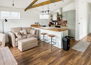 Ein modernes Wohnzimmer mit einer Couch, einem Hocker und zwei Barhocker. Ein Küchenbereich mit einer Holzarbeitsplatte und einem Herd ist zu sehen.