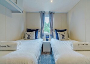 A twin bedroom with two beds, a nightstand, a lamp, and a window with curtains. - Premium 2 bed holiday home with hot tub Pet (Bassenthwaite, near Keswick)