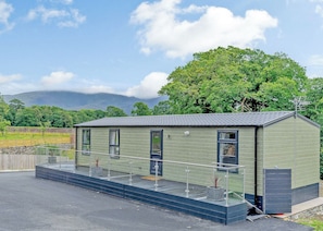 A small, single-story building with a sloped roof, a balcony with a glass railing, and a door. - Premium 2 bed holiday home with hot tub Pet (Bassenthwaite, near Keswick)