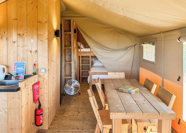 Una carpa con una cama litera, una mesa con sillas, un ventilador y un extintor.