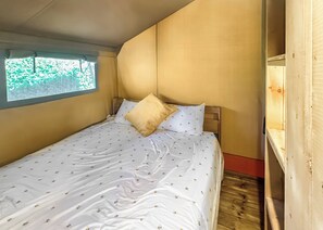 A single bed with a white sheet and a patterned pillow, a window with a view of greenery, and wooden walls. - Sea View Safari Tents (Hemsby)