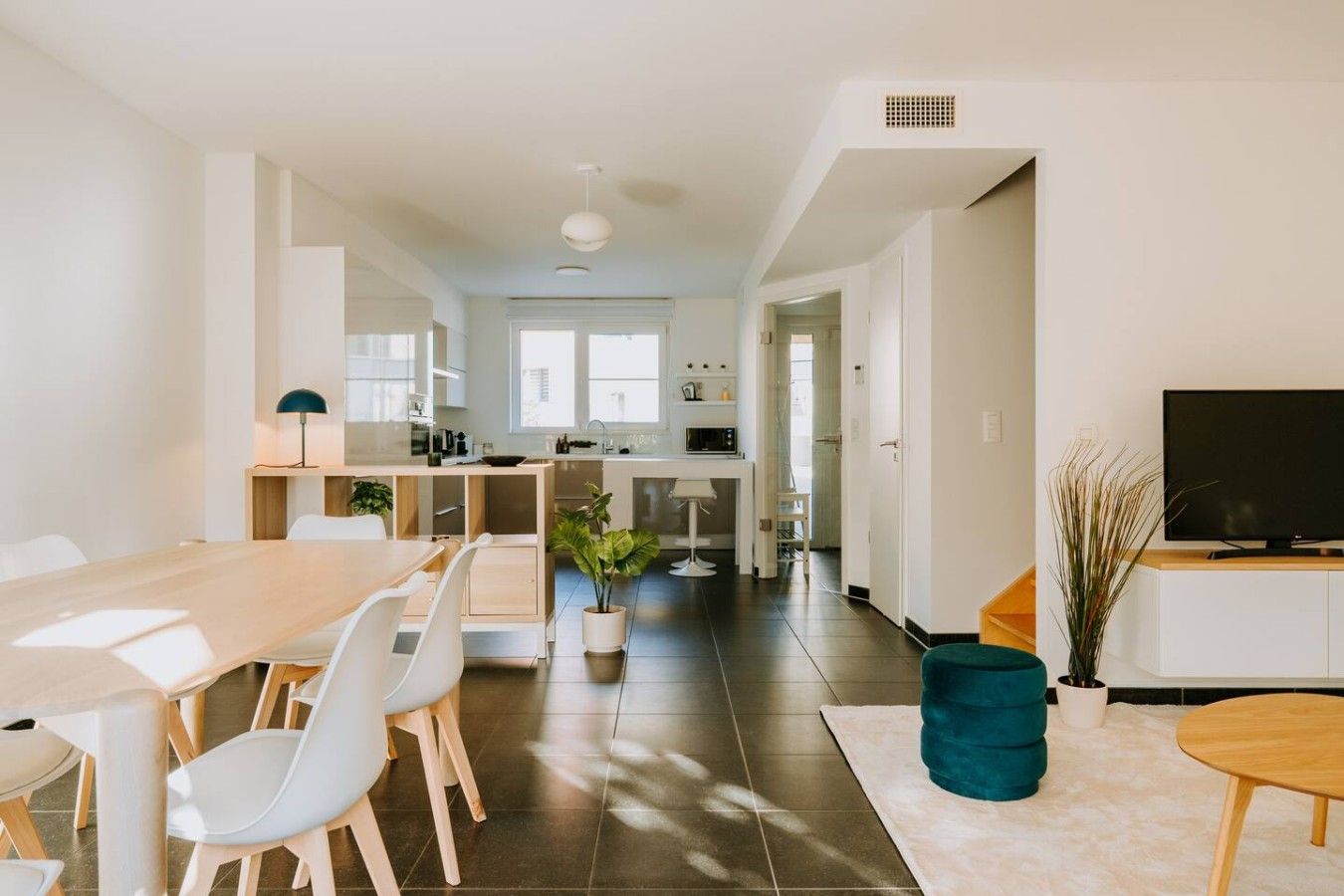 Une cuisine moderne avec des meubles blancs, un coin repas avec une table en bois et des chaises blanches, et un salon avec une télévision et une plante.