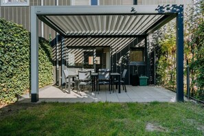 A covered patio area with a table and chairs, surrounded by greenery and a building. - 3-Bedroom House with Garden (Liège)