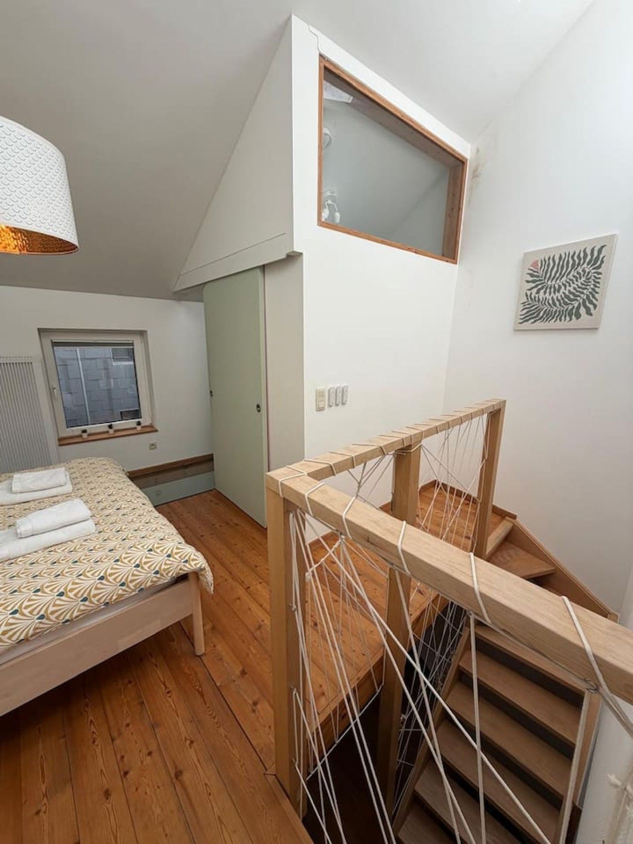 Un dormitorio con cama, ventana, escalera de madera y una obra de arte colgada en la pared.