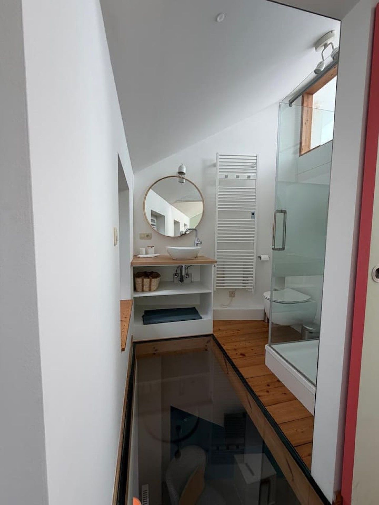 Un baño pequeño con cabina de ducha de vidrio, un espejo redondo y un lavamanos blanco con estante de madera.