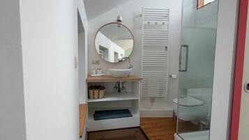 Un baño pequeño con cabina de ducha de vidrio, un espejo redondo y un lavamanos blanco con estante de madera.