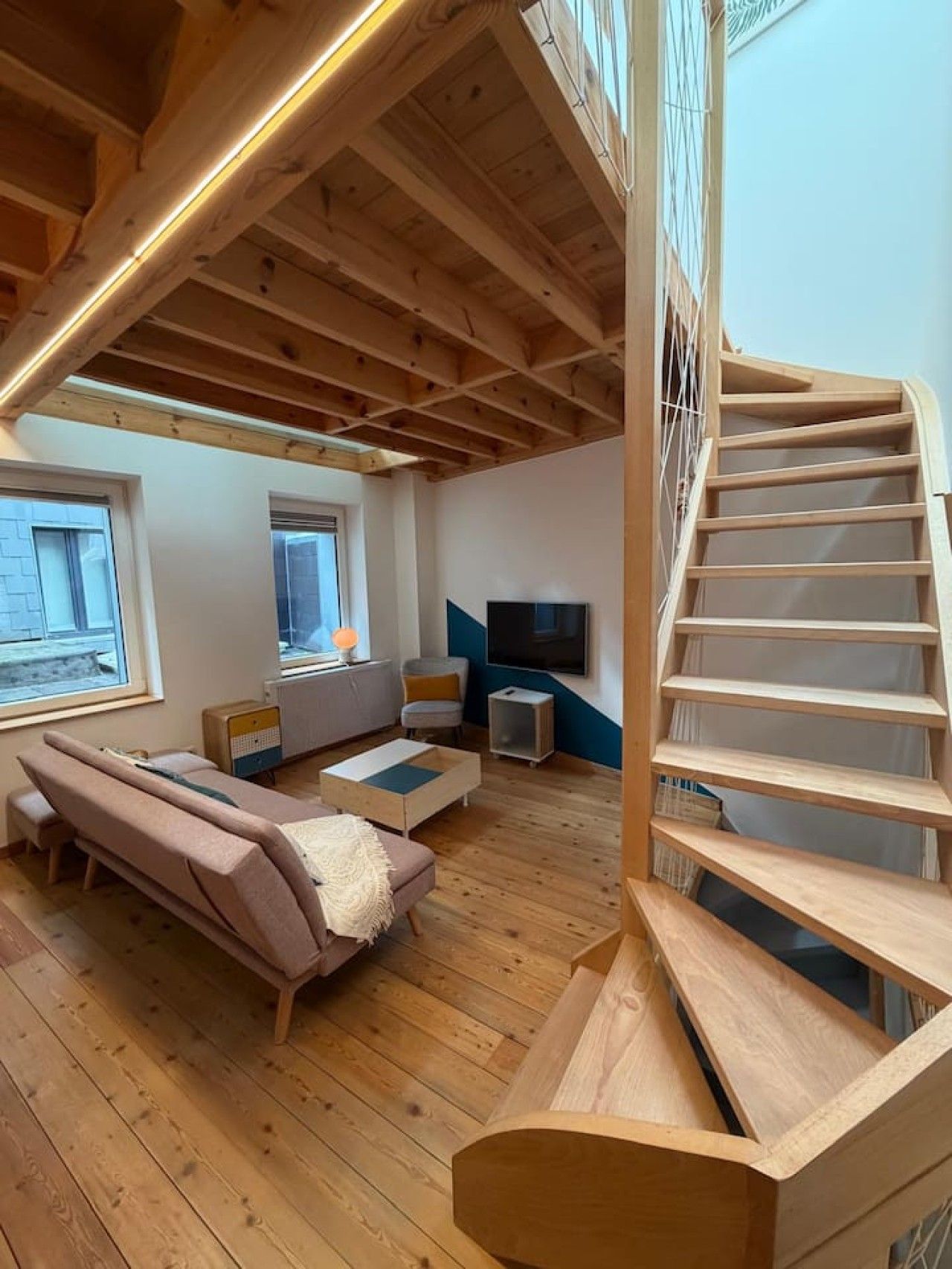 Een ruime loft met een houten wenteltrap, een bank, een televisie en een groot raam.