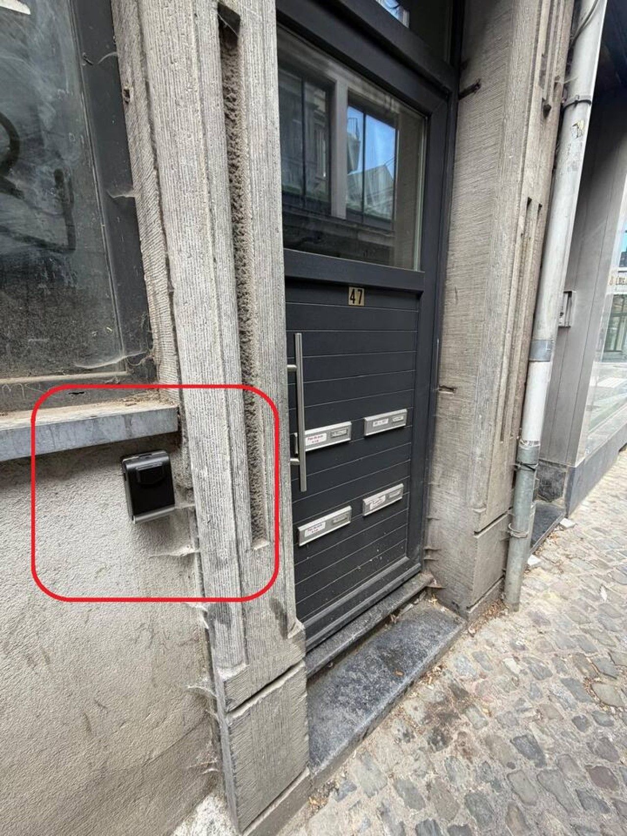 Une façade d’immeuble avec une porte de couleur sombre, une fenêtre au-dessus et une petite ouverture rectangulaire sur le côté gauche.