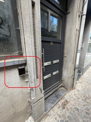 Une façade d’immeuble avec une porte de couleur sombre, une fenêtre au-dessus et une petite ouverture rectangulaire sur le côté gauche.