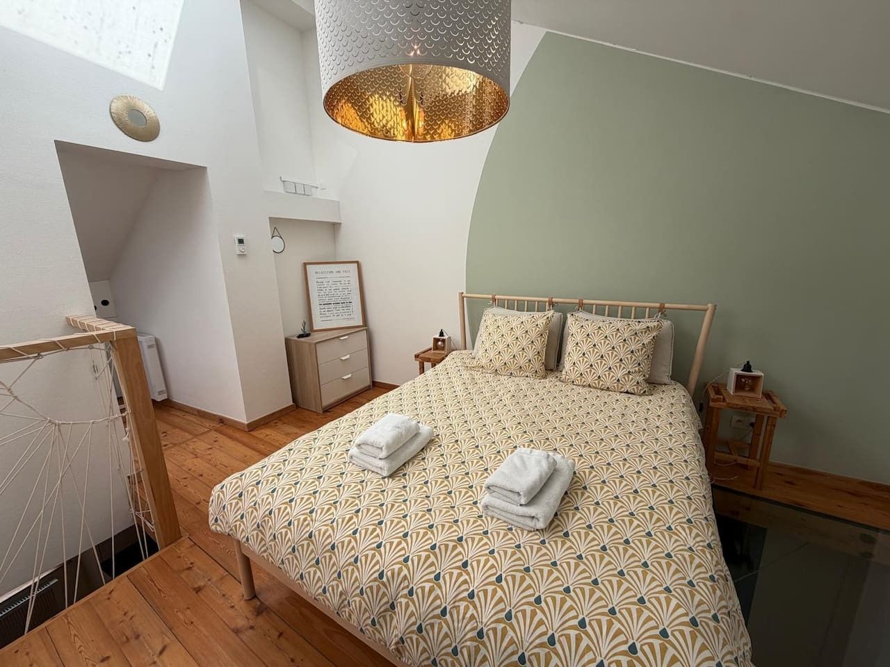 A bedroom with a patterned bedspread, a hanging light fixture, a bedside table with a lamp, and a small framed picture on the wall.