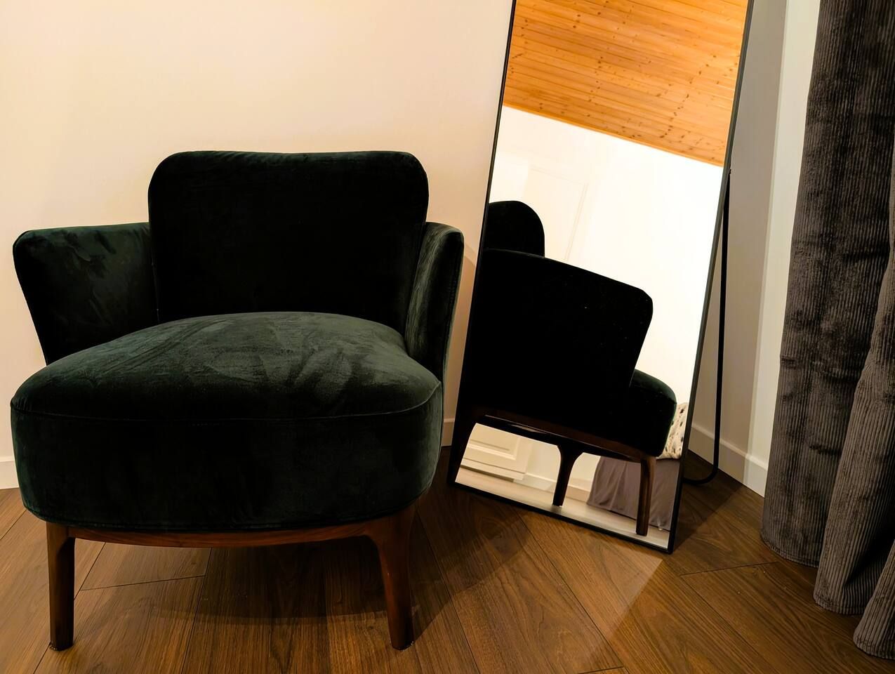 A dark green armchair with wooden legs, positioned next to a mirror reflecting a room with a wooden ceiling.