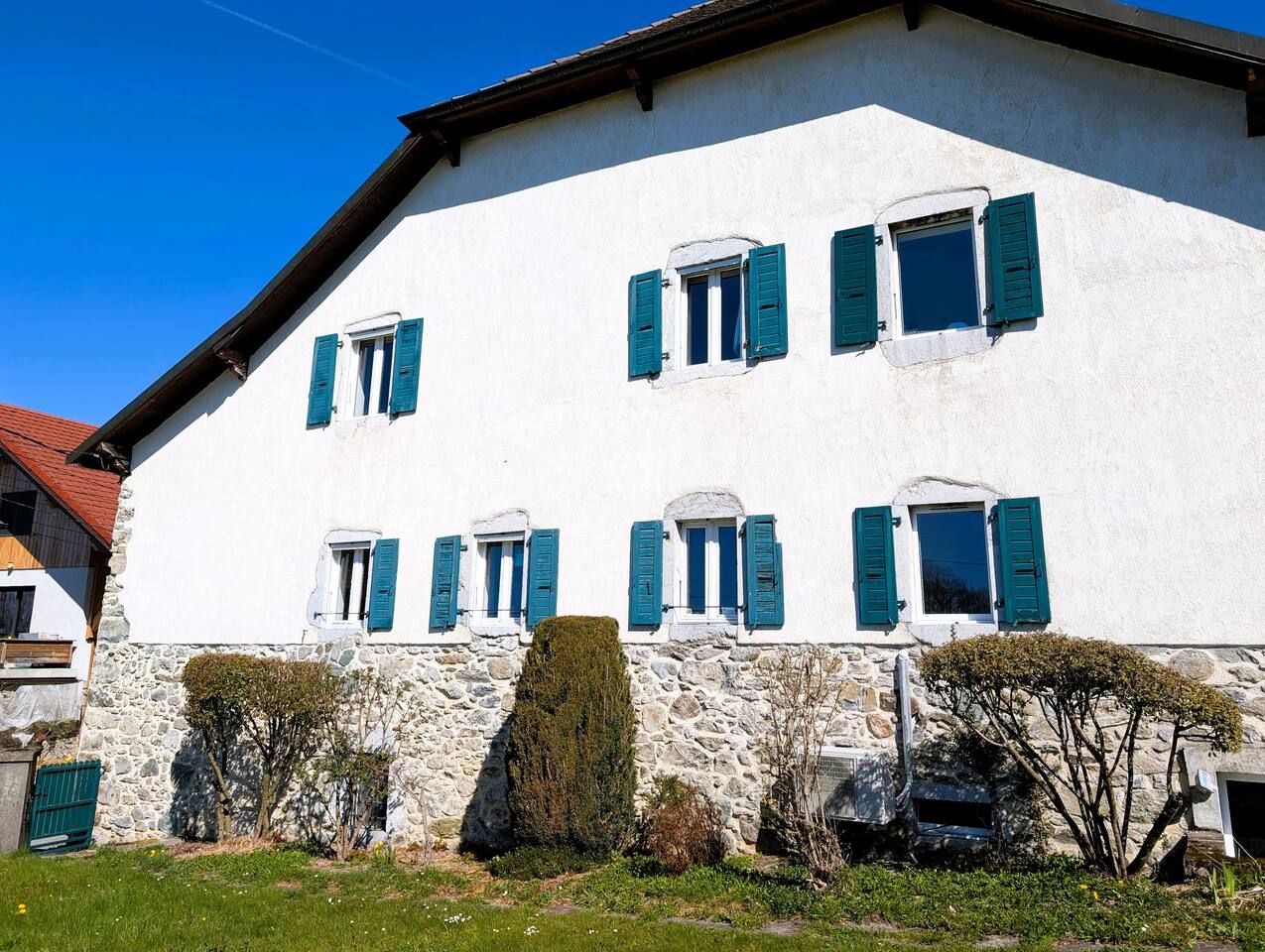 A white building with multiple windows and green shutters, surrounded by stone walls and greenery.