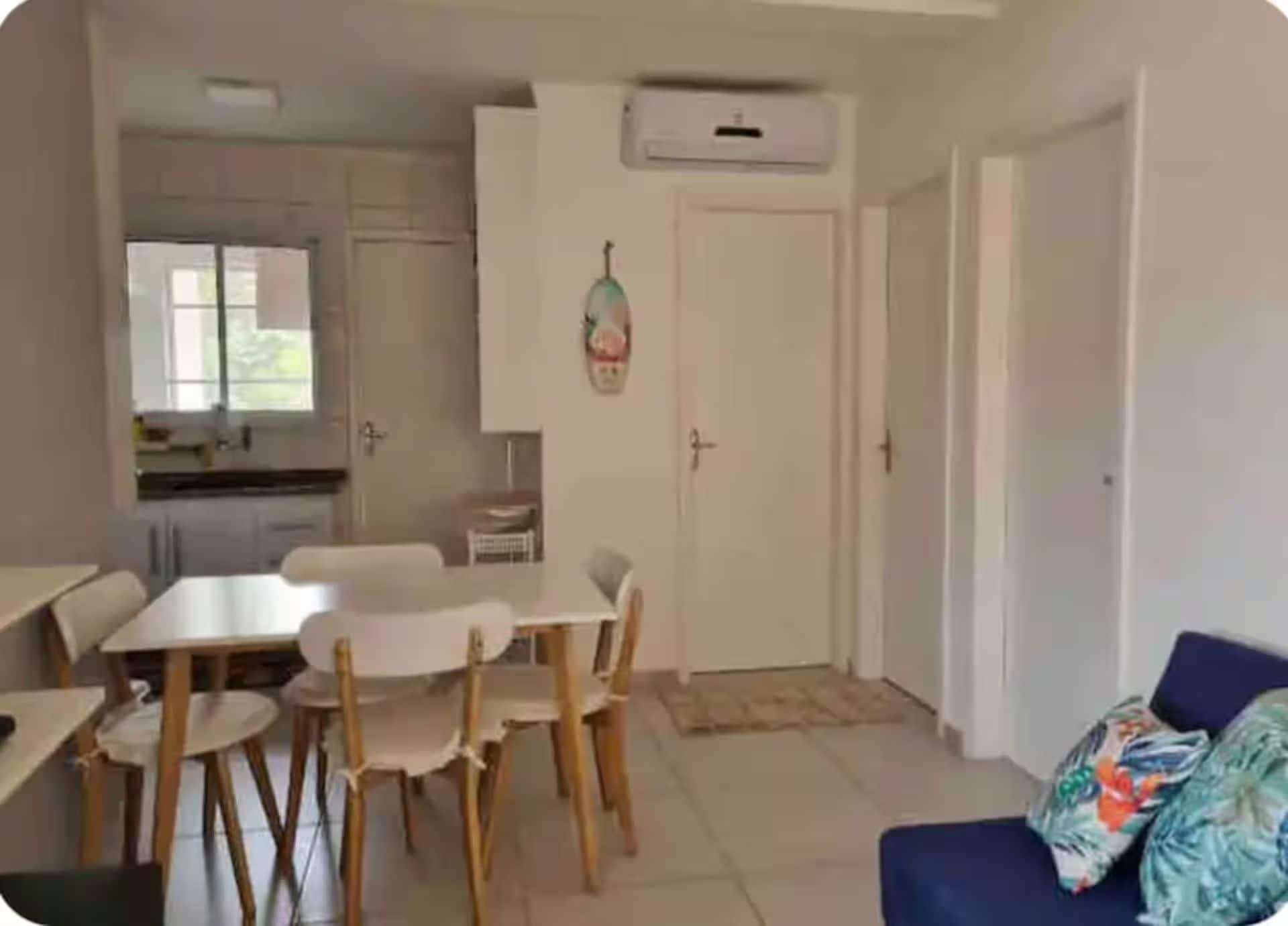 A room with a dining table and chairs, a sofa, a kitchen area with cabinets, and an air conditioning unit.
