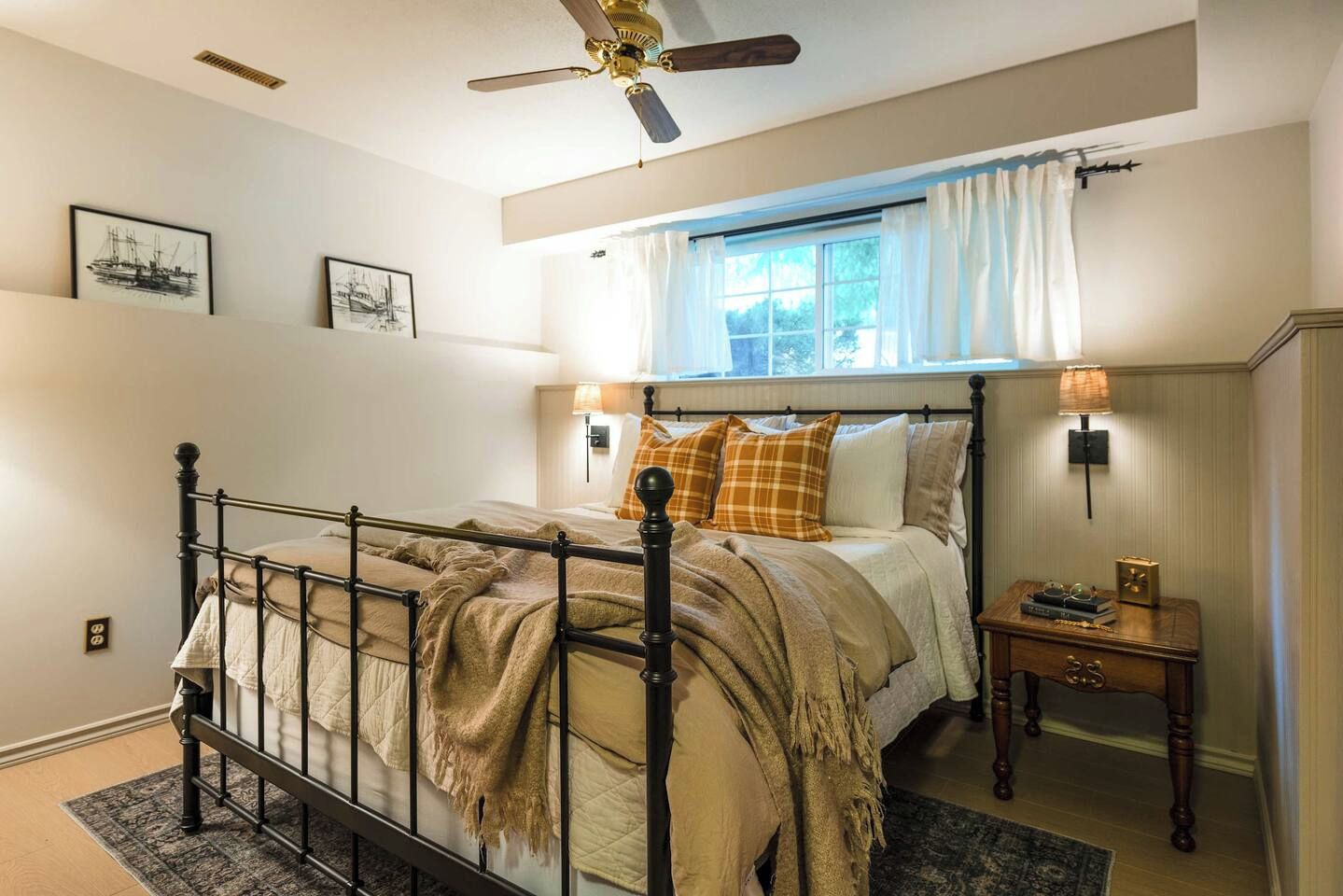 Une chambre à coucher comprenant un lit, une table de chevet, une fenêtre avec des rideaux, un ventilateur de plafond et des lampes fixées au mur.