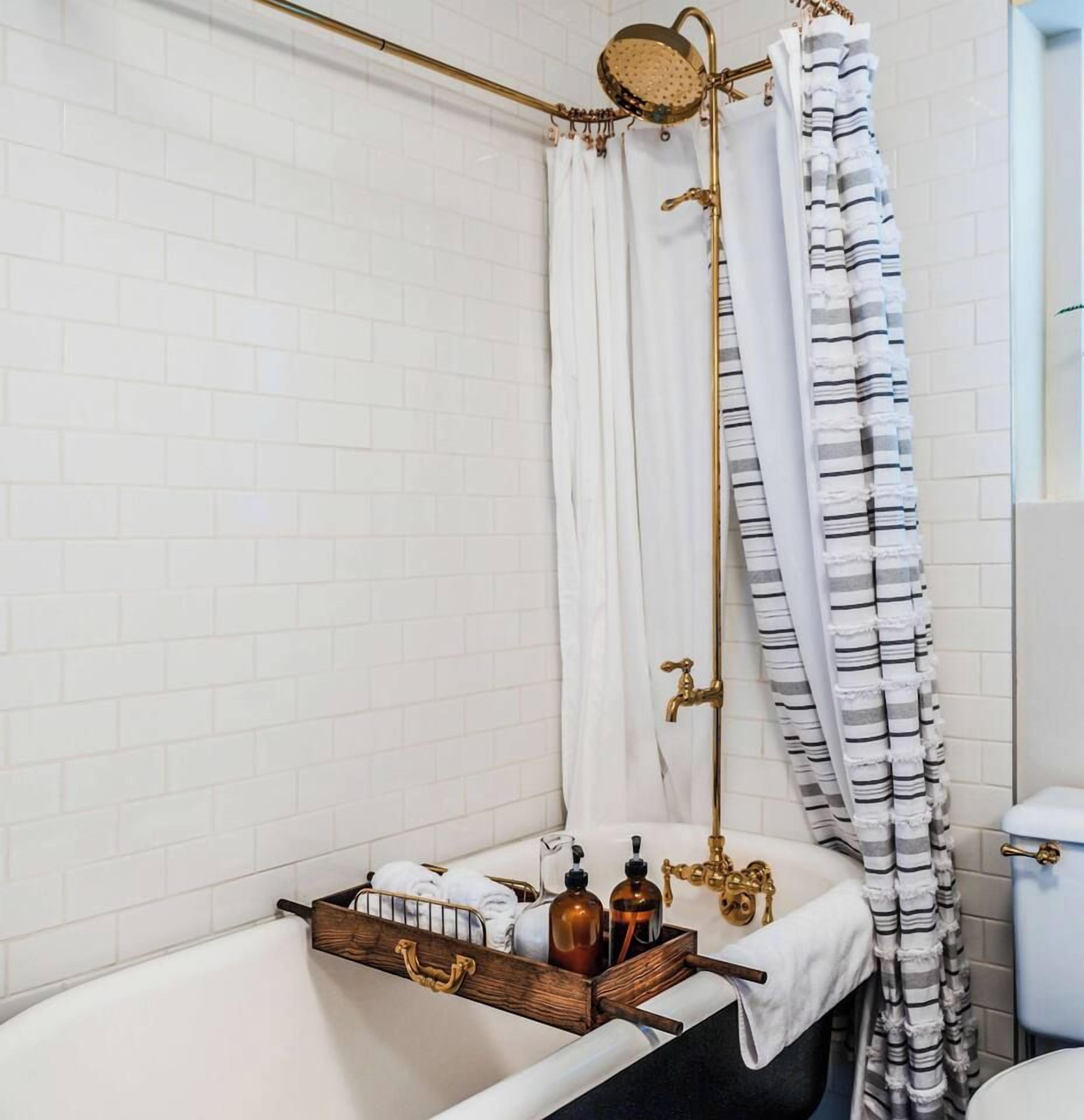 A bathroom with a white bathtub, a shower with a gold showerhead, and a striped shower curtain. There is a wooden tray with toiletries on the edge of the bathtub.