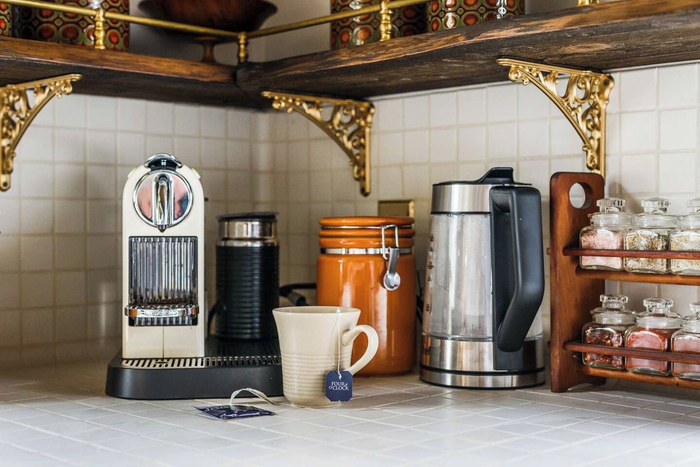 Keittiössä on vanhanaikainen espressokone, kahvipannu, muki, termospullo ja maustepurkkeja hyllyllä.