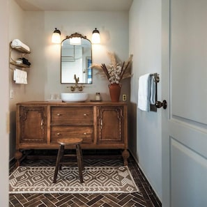 Une salle de bain avec un meuble-lavabo en bois, un grand miroir, deux appliques murales, un porte-serviettes et un tapis à motifs.