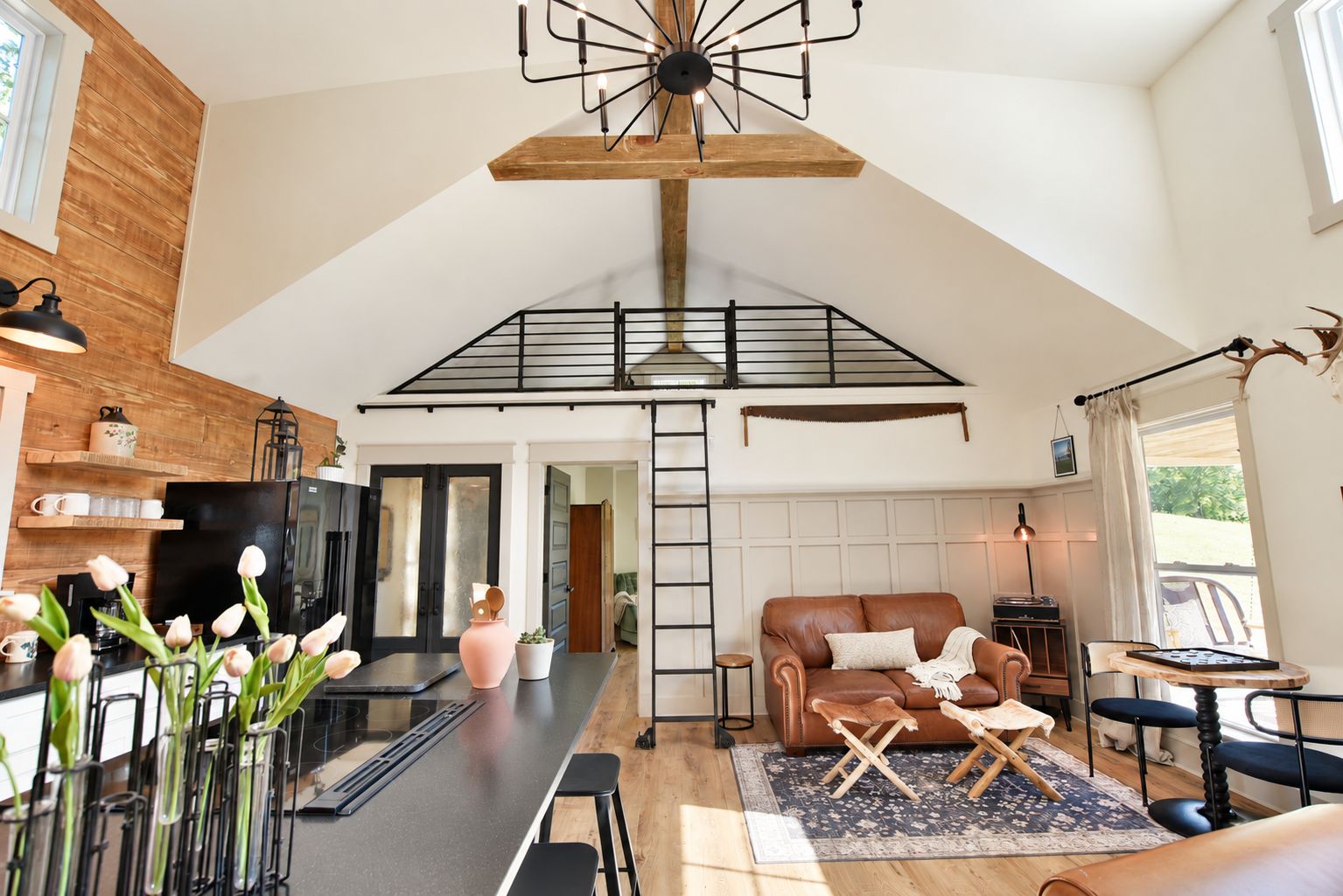 A spacious living area with a high ceiling, a loft bedroom with a wooden beam, a kitchen with modern appliances, and a cozy seating area with a leather sofa and a small table.