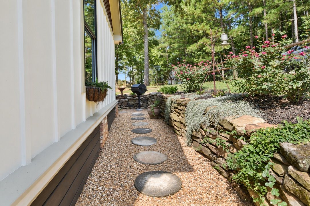 A pathway with stepping stones leading to a house with a stone wall and a garden with plants and flowers.