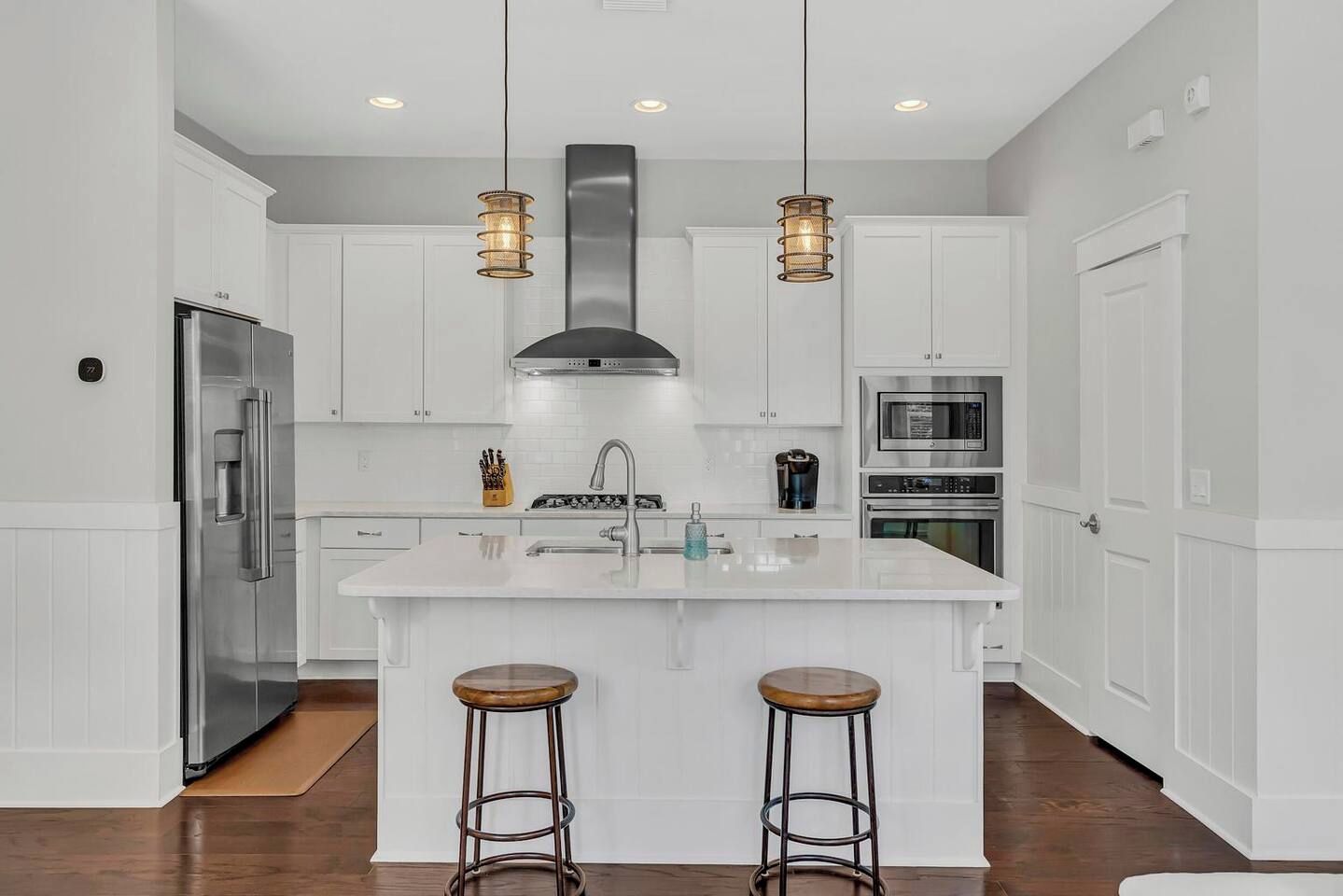 A modern kitchen with white cabinetry, stainless steel appliances, a central island with stools, pendant lighting, and a visible water dispenser.