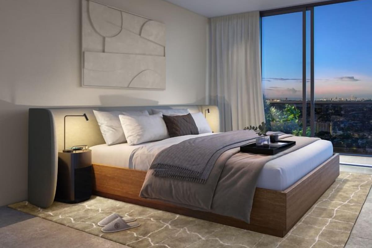 Une chambre moderne avec un grand lit, une table de chevet, une lampe et une vue sur le paysage urbain grâce à de grandes fenêtres.