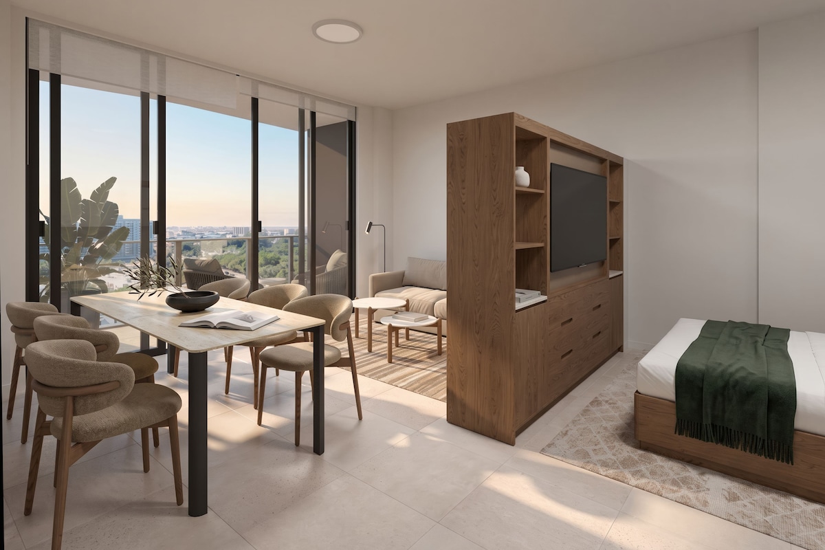 Une chambre moderne avec une grande fenêtre, un lit recouvert d’une couverture verte, un meuble TV en bois et un coin repas avec des chaises et une table.