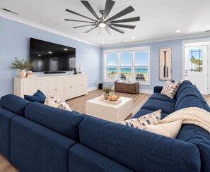 A living room with a sectional sofa, a flat-screen TV, a ceiling fan, and a view of the ocean through large windows. - NEW! Ocean Views-Steps to Beach-Pool+Spa-Luxury 8BR (Atlantic Beach)