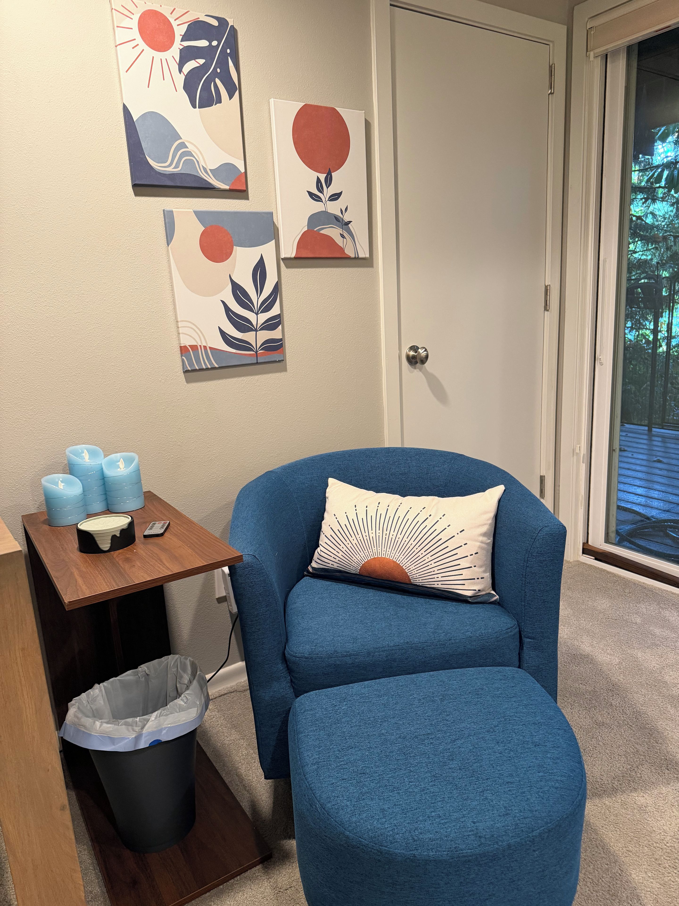 Une pièce avec un fauteuil bleu, une petite table en bois avec des bougies, une poubelle et des œuvres d’art murales.