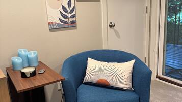Une pièce avec un fauteuil bleu, une petite table en bois avec des bougies, une poubelle et des œuvres d’art murales.
