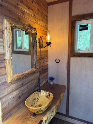 A rustic bathroom with wooden walls, a wooden basin, a wall-mounted light, and a window with a sign. - Cowboy-Themed Tent Getaway (Honey Grove)
