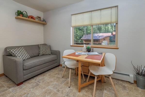 A living room with a grey sofa, a wooden table, and chairs. There is a window with blinds, a shelf with decorative items, and a potted plant.
