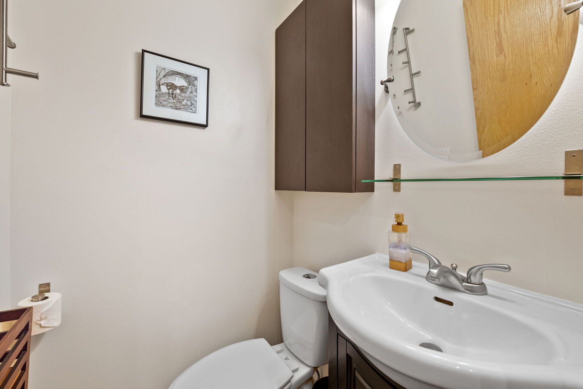 Ein Badezimmer mit einem weißen Waschbecken, einer Toilette, einem gerahmten Bild an der Wand und einem Spiegel mit Holzrahmen.