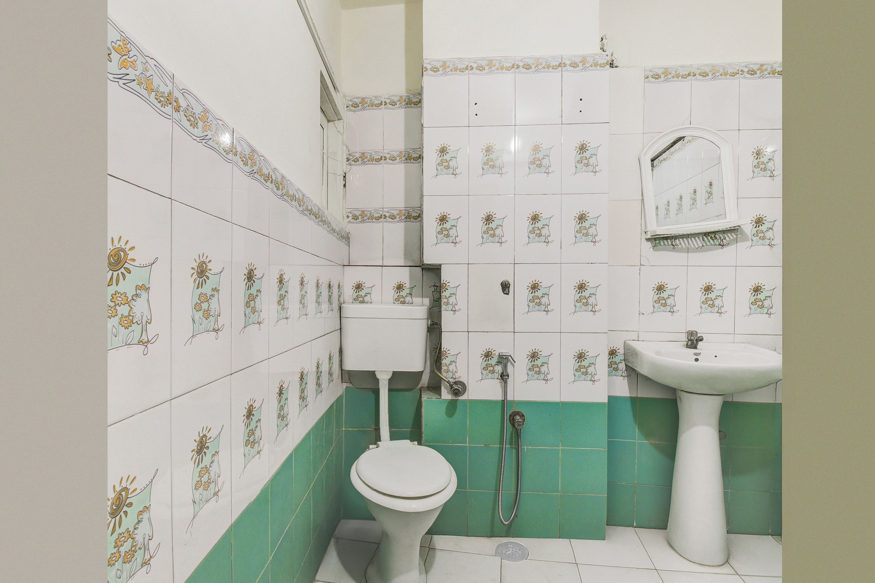 A bathroom with white tiles featuring floral patterns, a white toilet, a green-tiled shower area, and a white pedestal sink.