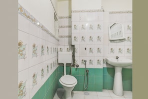 Un baño con azulejos blancos con diseños florales, un inodoro blanco, una zona de ducha con azulejos verdes y un lavabo blanco sobre pedestal.