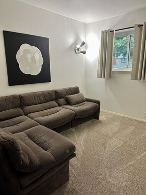 Un salon avec un canapé d’angle, un luminaire fixé au mur et une fenêtre avec des rideaux.