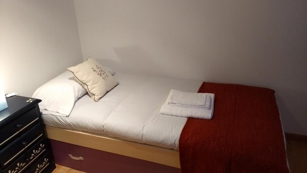 A single bed with a red blanket and a white pillow, a wooden dresser with drawers, and a white wall.