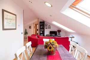 Un comedor con una mesa dispuesta para cuatro, un sofá rojo al fondo y un tragaluz arriba.