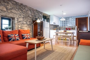 Una sala de estar con pared de piedra, un sofá con cojines, una mesa de madera y una zona de cocina con una mesa de comedor y sillas.